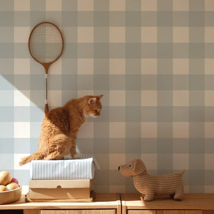 Cat sitting on a box with a badminton racket against a checkered wall, next to a dog figurine.