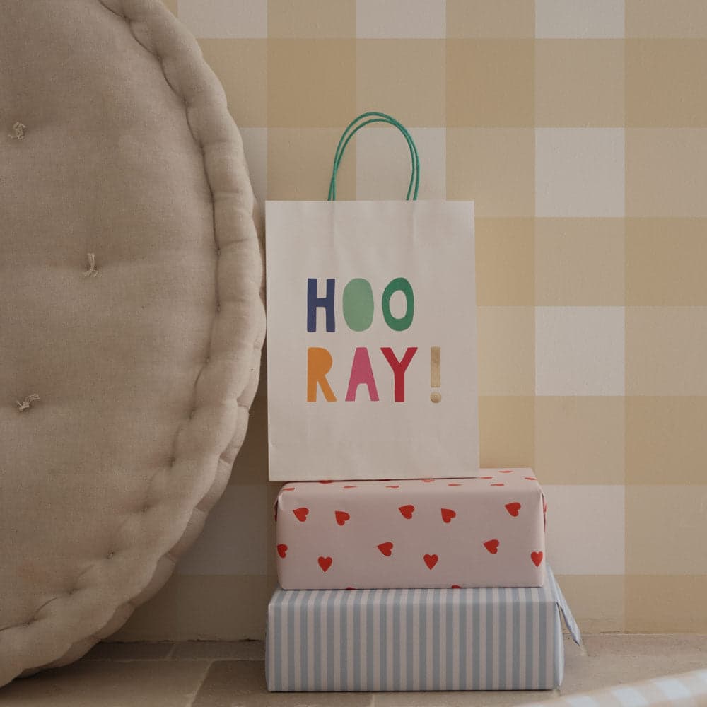 Stack of wrapped presents with a 'Hooray!' bag against a checkered wall.