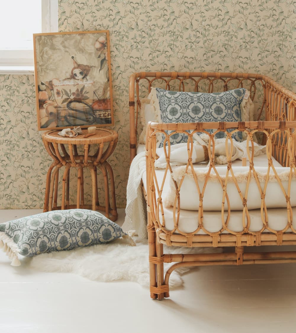 Rattan crib with white bedding and patterned pillows in a room with floral wallpaper.